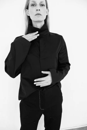 Black and white portrait of an adult woman in a high-collared black shirt and black pants against a white wall, fashion editorial.