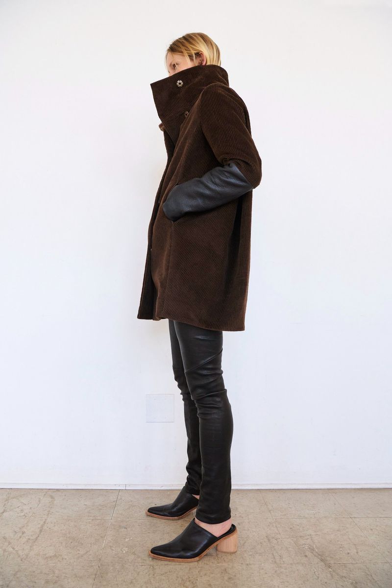 Fashion model wearing a brown coat and black pants stands against a white background in a studio, showcasing a stylish outfit.