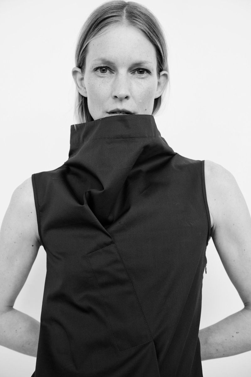 Monochrome portrait of a fair-skinned adult woman with fair hair wearing a sleeveless dress with a high, draped collar against a white background.