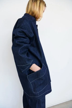 Blonde person wearing a dark blue denim jacket with white stitching against a white background, standing indoors, with a hand in their pocket.