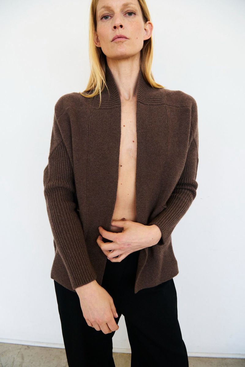 Adult woman poses in a photo studio wearing an open brown cardigan and black pants against a neutral white backdrop, fashion portrait.