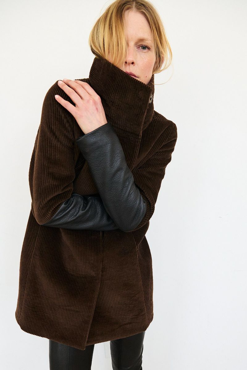 A woman in a corduroy jacket with leather sleeves poses in a studio against a white background, looking directly at the camera.