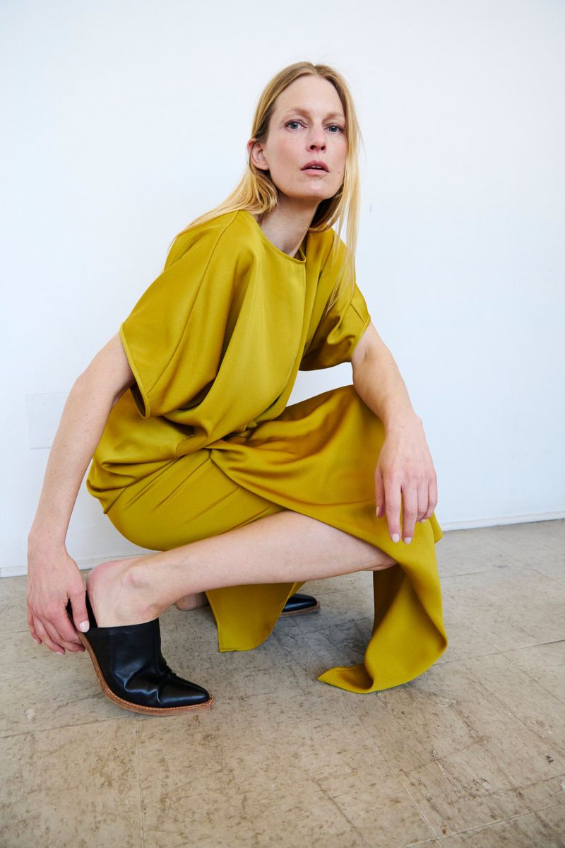 Woman poses in a mustard-yellow dress and black heeled mules against a stark white wall, showcased in a fashion-forward studio shot.