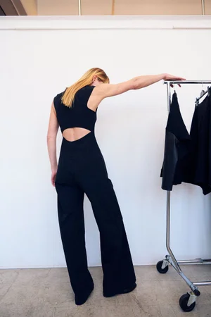 Model seen from behind wearing a black cut-out jumpsuit leaning against a rolling clothing rack in a bright white studio.