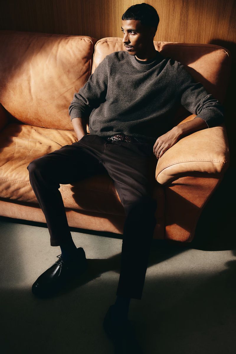A young adult male with dark hair sits casually on a tan leather couch in a dimly lit indoor space wearing a grey sweater.