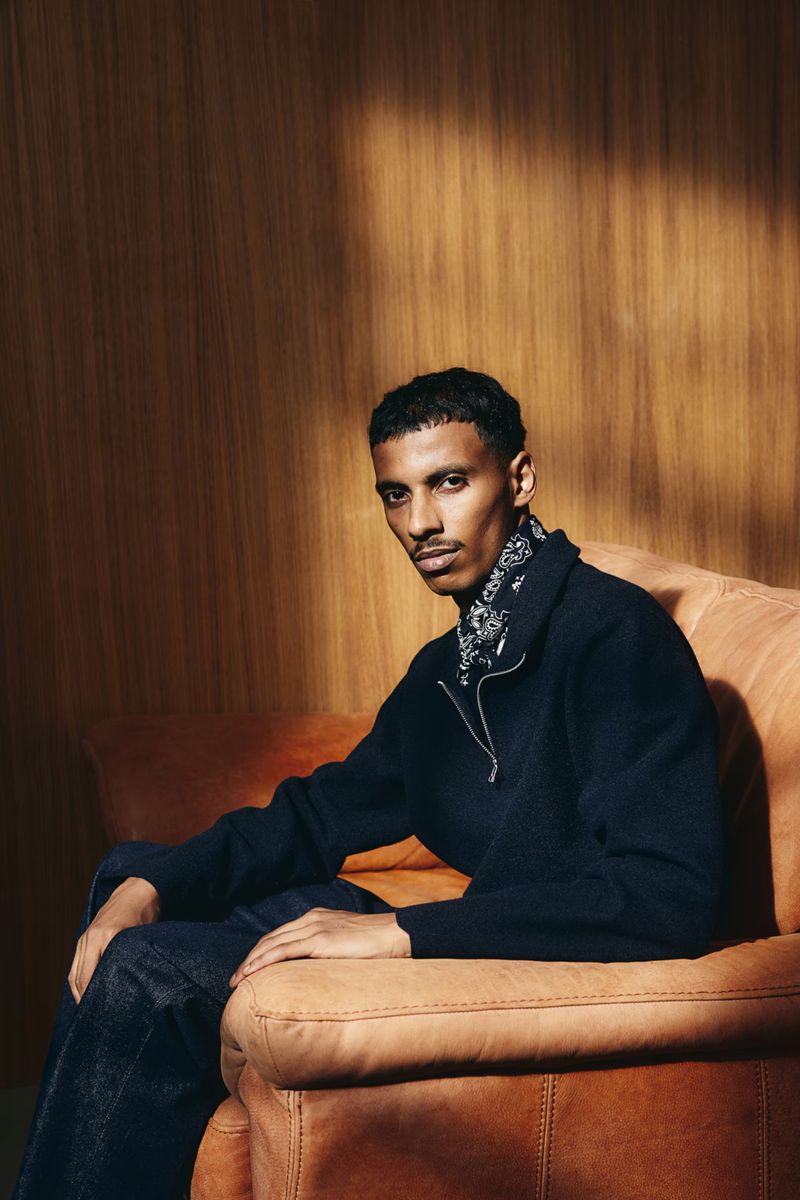 An adult male sits on a brown leather sofa indoors. He is wearing a navy-blue sweater, dark blue jeans, and a black-and-white patterned bandana.