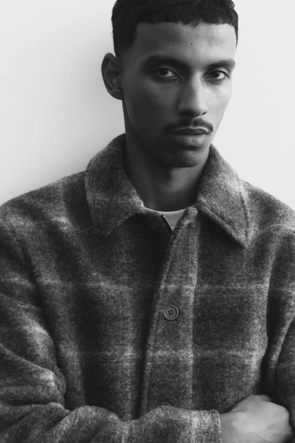A stylish adult male with dark hair and a mustache wears a checkered coat indoors against a white wall, captured in a black and white portrait.