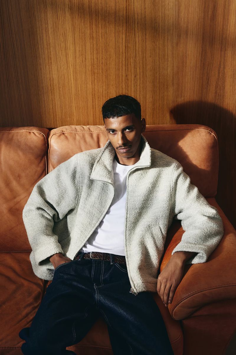 A man sits casually on a leather sofa in a naturally lit indoor setting, wearing a white shirt, fuzzy jacket, and dark jeans.