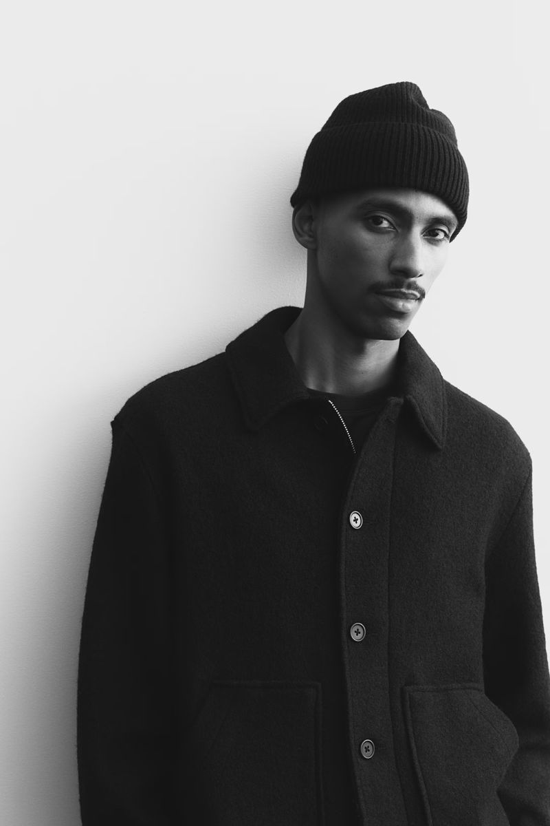 Black and white portrait of a young adult man in a dark jacket and beanie against a light wall, captured in a medium studio shot.