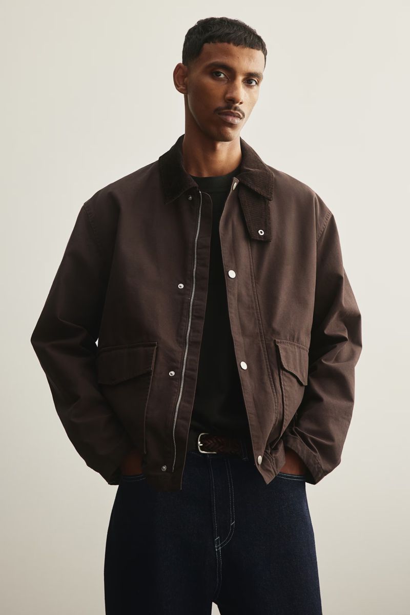 Dark-haired adult male wearing a brown jacket, black shirt, and dark blue jeans poses in a studio with a neutral background.