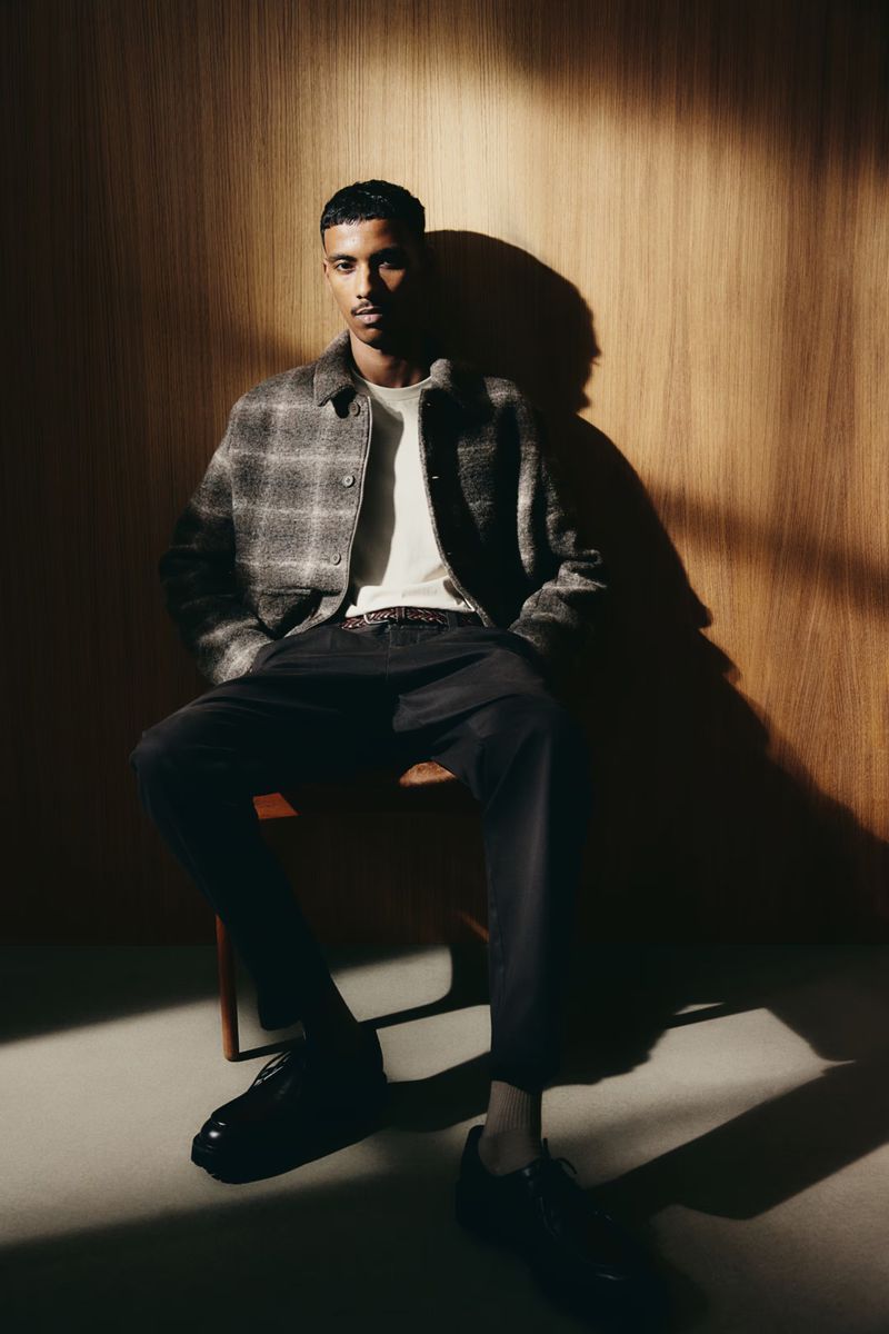 Model Mohamed Hassan seated on a wooden chair wearing a plaid jacket against a wood panel wall