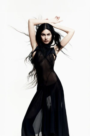 Fashionable woman with long dark hair poses in a sheer black dress against a white backdrop in a studio shot for an editorial fashion layout.