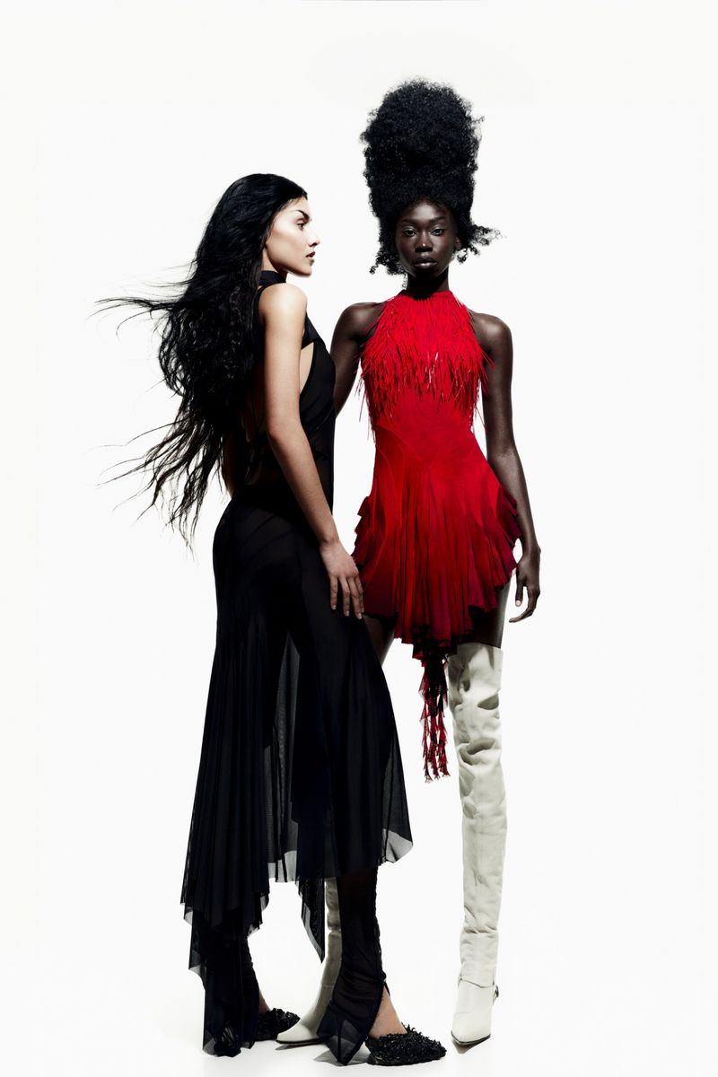 Two women pose in a fashion studio wearing elegant dresses, one red and one black, against a bright white backdrop in a vertical shot.