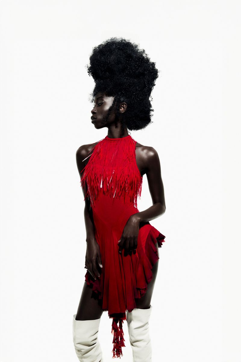 Black woman with voluminous afro wearing a red fringed dress and white knee-high boots against a plain white background