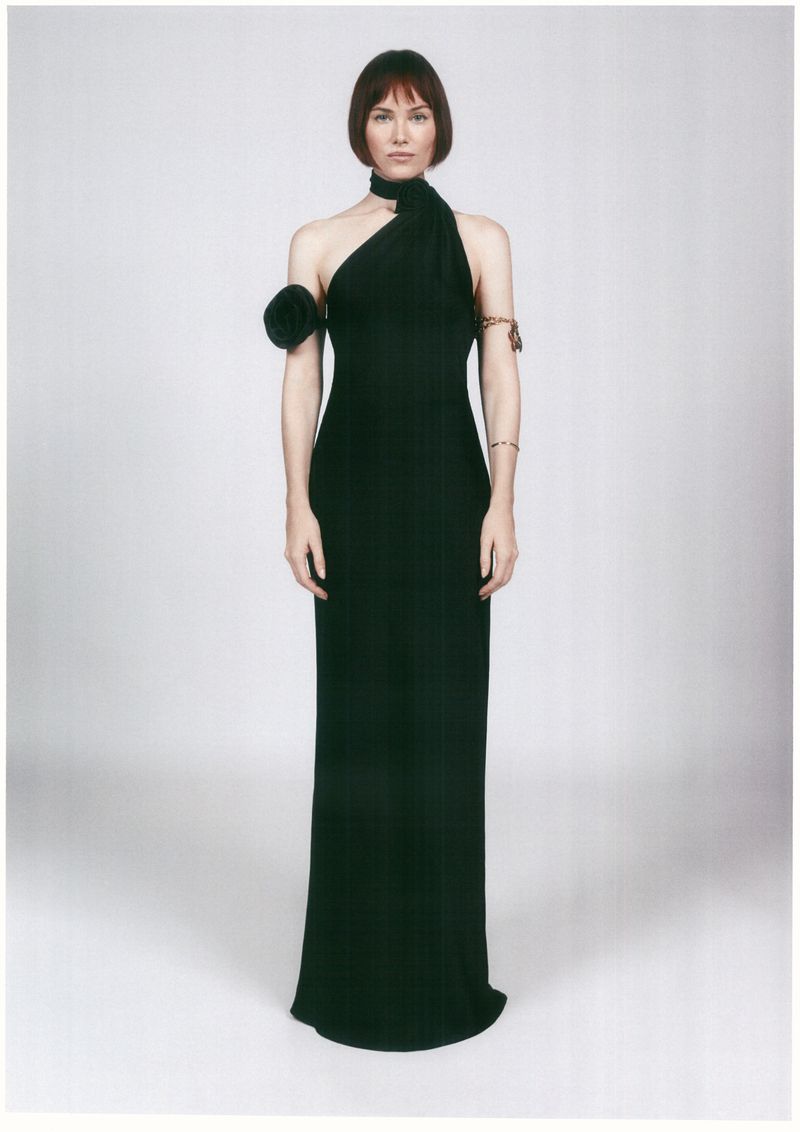 An adult woman is posing in a full shot, wearing a black gown with a neck wrap and shoulder detail against a light neutral backdrop.