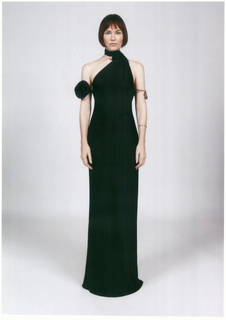 An adult woman is posing in a full shot, wearing a black gown with a neck wrap and shoulder detail against a light neutral backdrop.