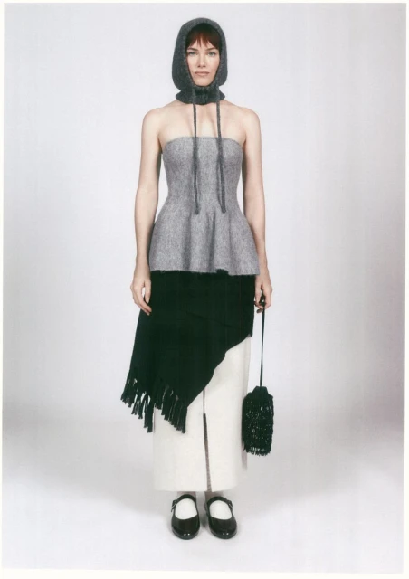 Woman in gray knit hood and top, black draped shawl, white skirt with slit, black Mary Jane shoes, and beaded bag on white.