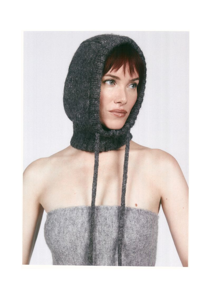 A woman is shown in a gray knit hood and top, posing in a studio. The shot is close-up, highlighting the texture of the knitwear.