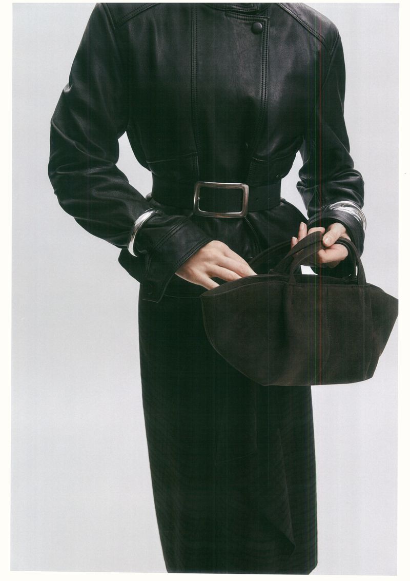 Fashion shot of a woman in a black leather coat with a belt, holding a small black purse against a plain white background.
