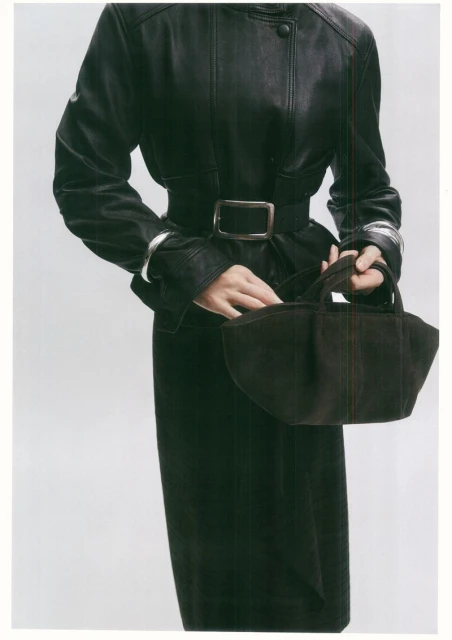 Fashion shot of a woman in a black leather coat with a belt, holding a small black purse against a plain white background.
