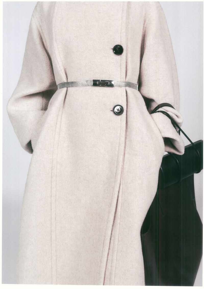 Close-up of a fair-skinned model wearing a light beige wool coat with black buttons and a silver belt against a white backdrop.