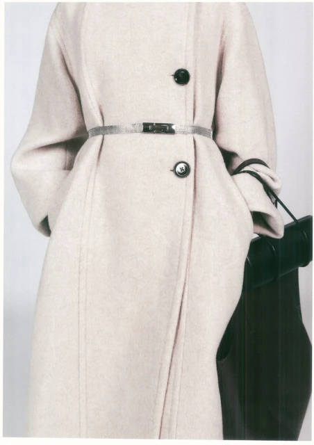 Close-up of a fair-skinned model wearing a light beige wool coat with black buttons and a silver belt against a white backdrop.