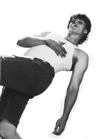 Monochrome portrait of a young adult man lying down wearing a white tank top and shorts in a light, bright indoor setting.