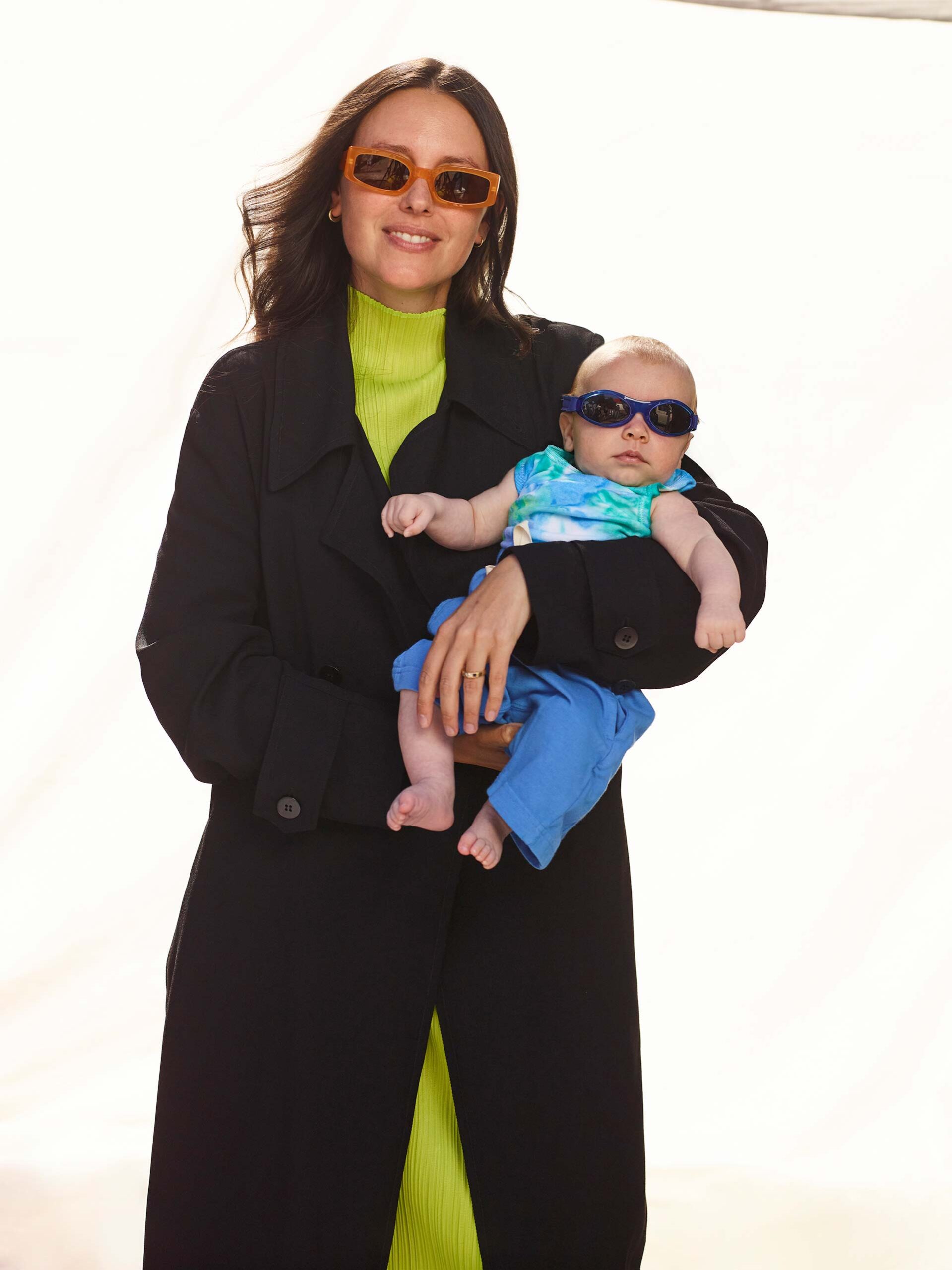 Stylish mother and baby pose outdoors wearing sunglasses and fashionable clothing in bright lighting against a neutral backdrop.
