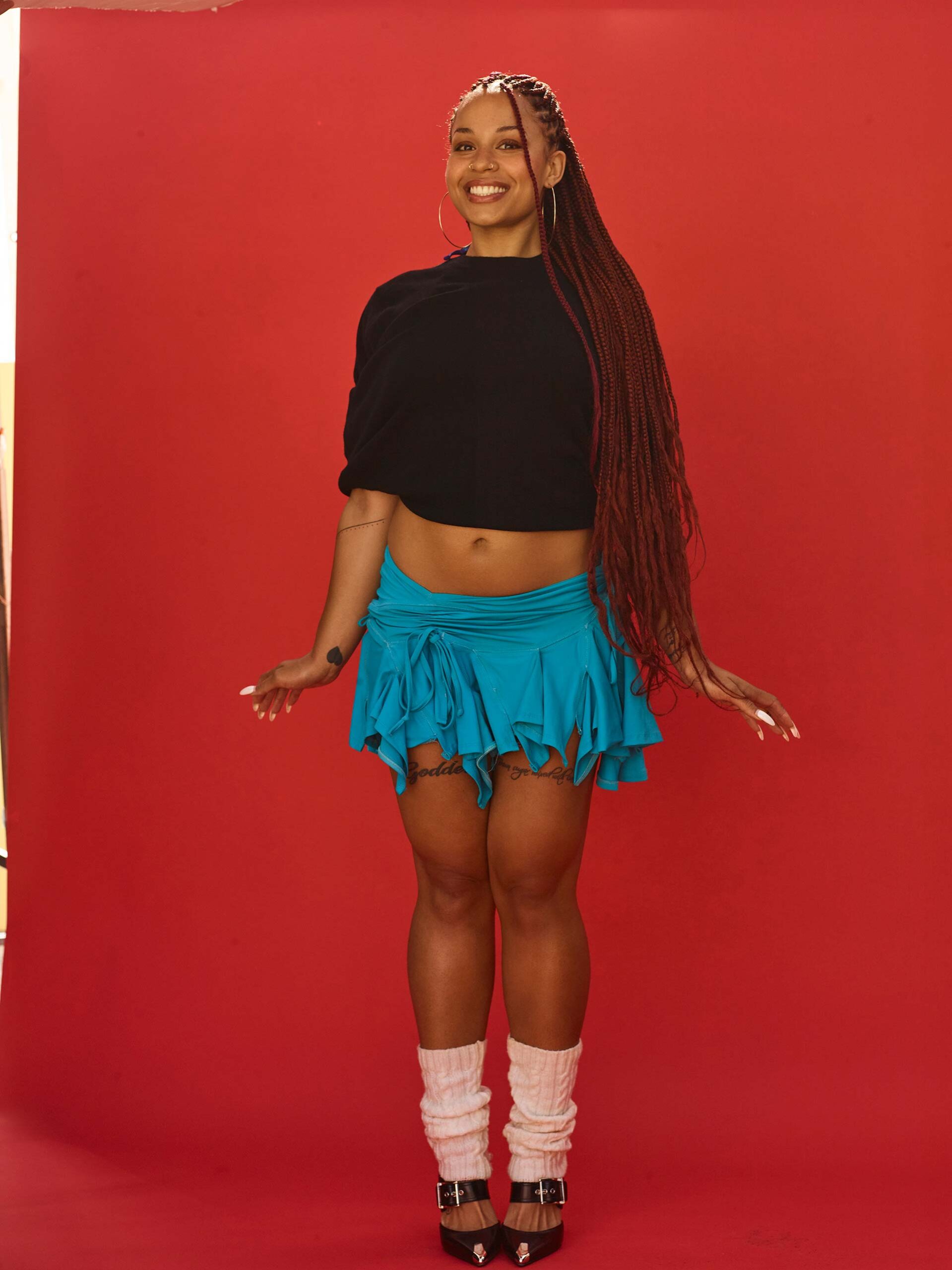 Full-length portrait of a young woman smiling in a blue skirt and black top, with long braids against a red background.