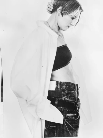 Black and white studio shot of a woman wearing a bra top, jeans, and a partially open button-down shirt, with her hand touching her hair.