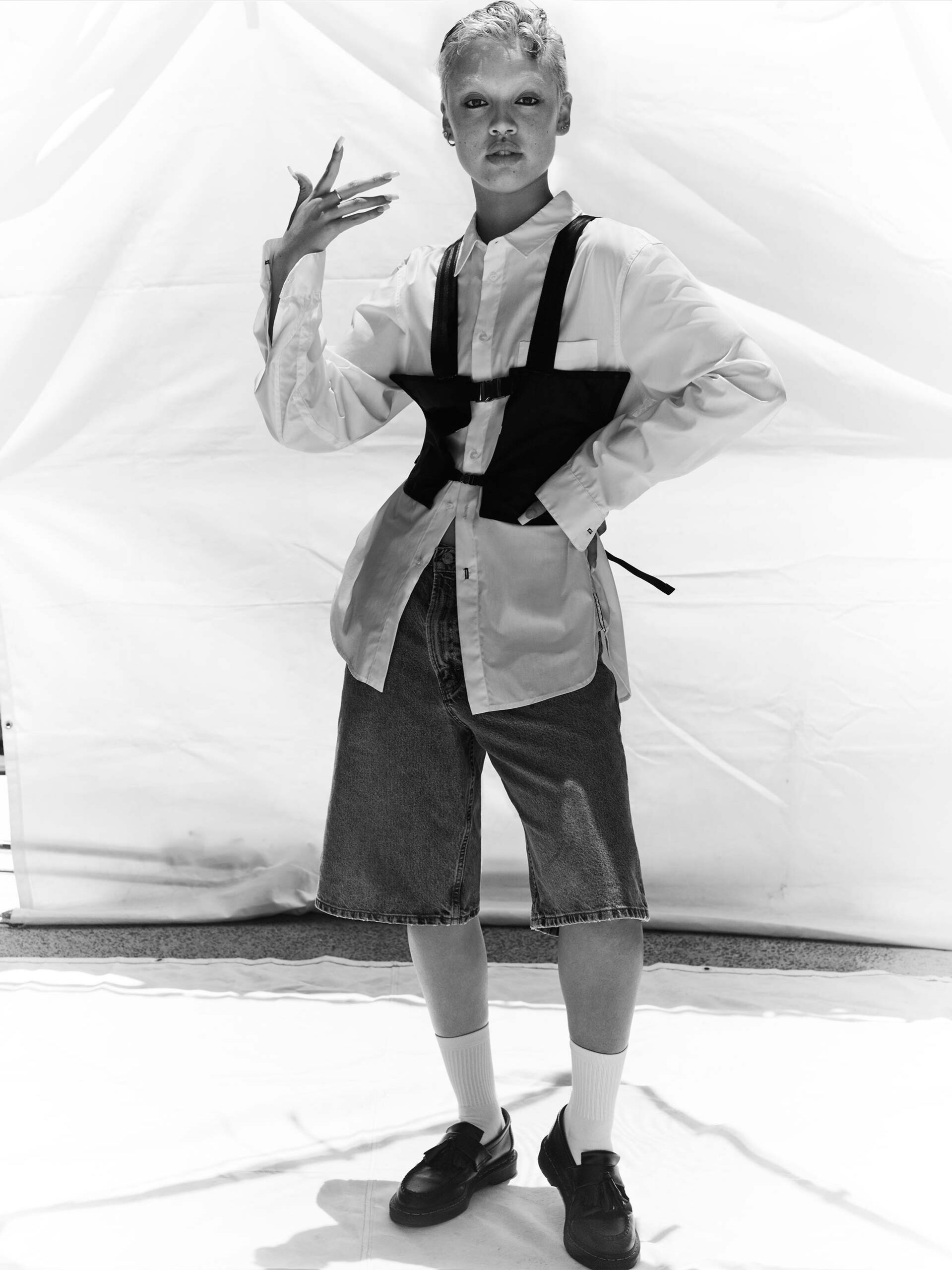 A model with short blonde hair wears a black corset, oversized white shirt, and denim shorts against a white backdrop.