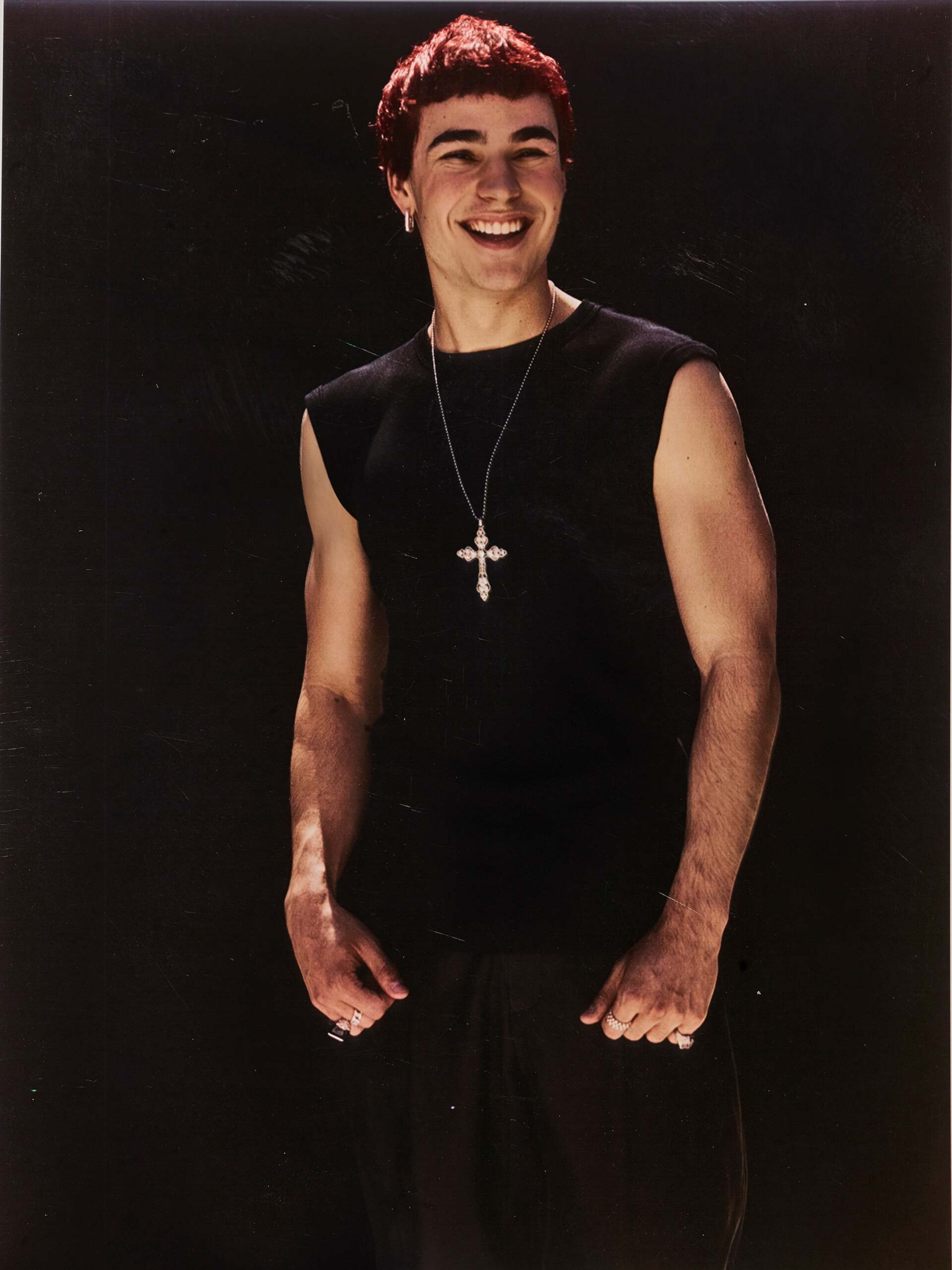 Portrait of a smiling adult male with dark hair, wearing a black tank top and silver cross necklace against a black background.