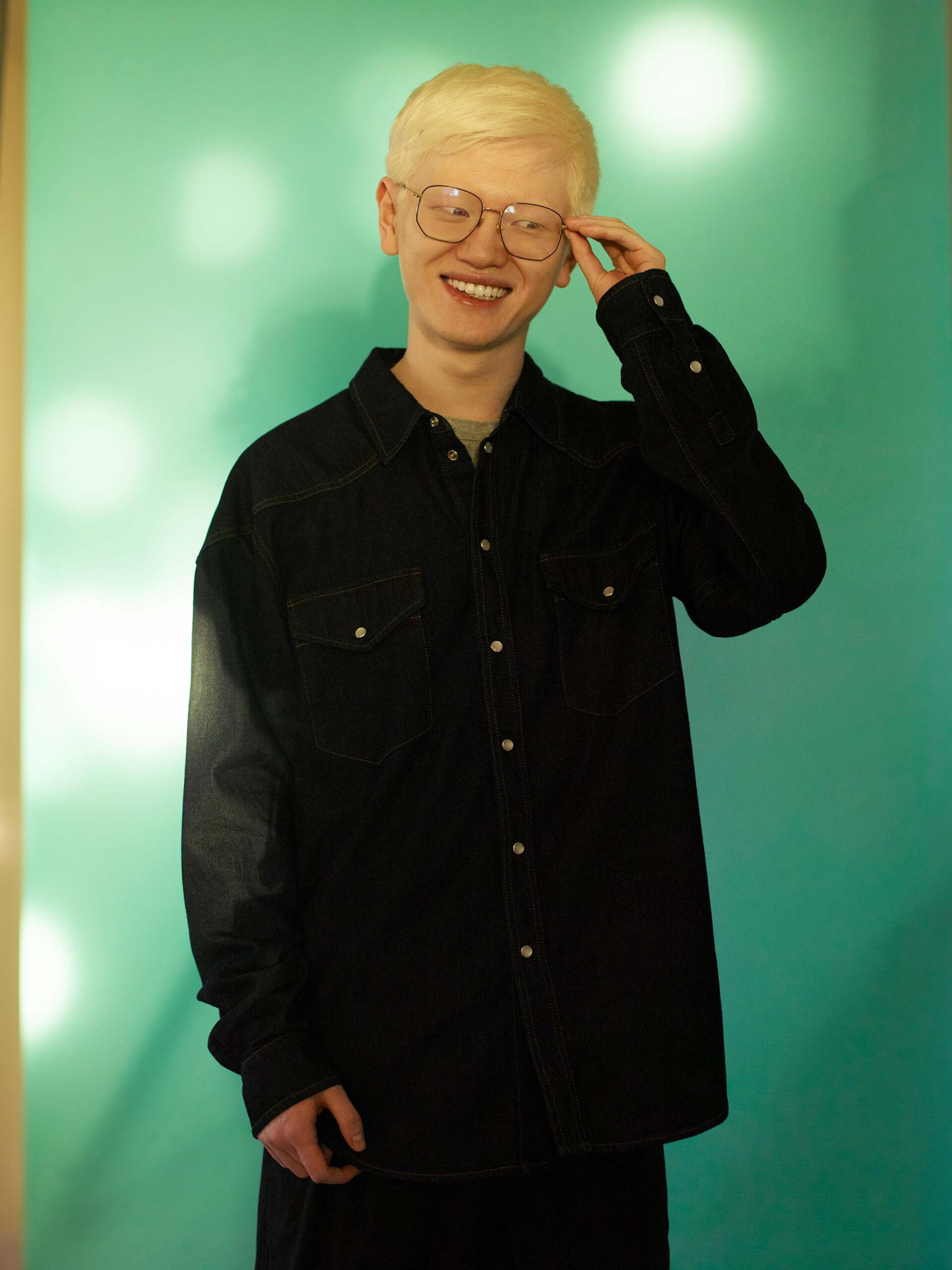 Smiling albino man in a dark denim shirt and glasses stands in front of a teal background in a medium portrait shot.