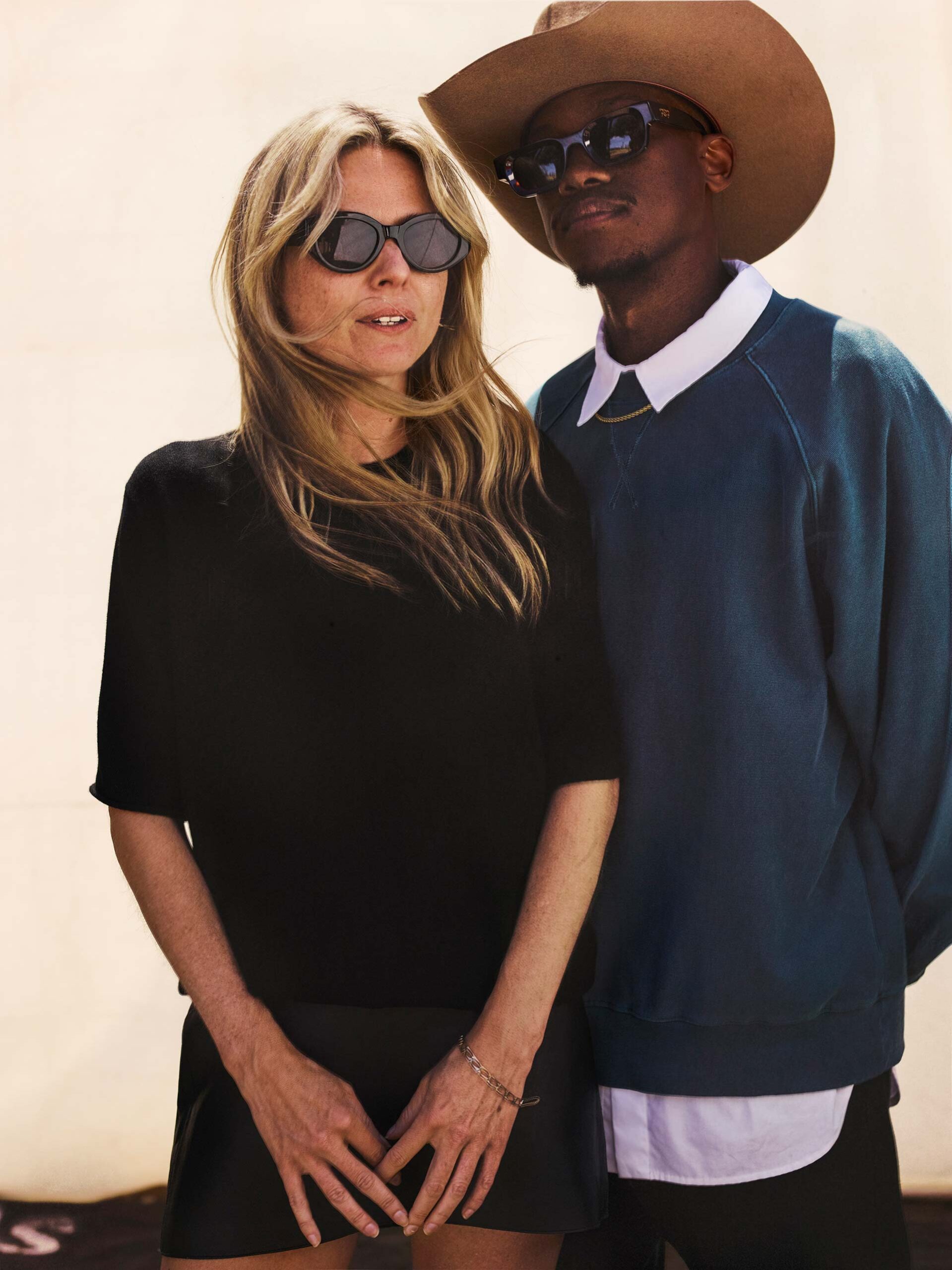 Editorial fashion shot of a Black adult male in a cowboy hat and a White adult female wearing sunglasses, standing indoors against a light wall.