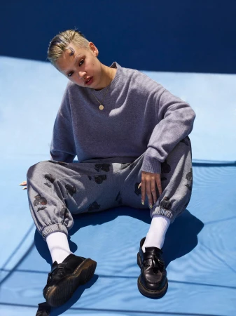 Model with short buzzcut hair wearing a grey sweater and patterned trousers seated on a blue surface