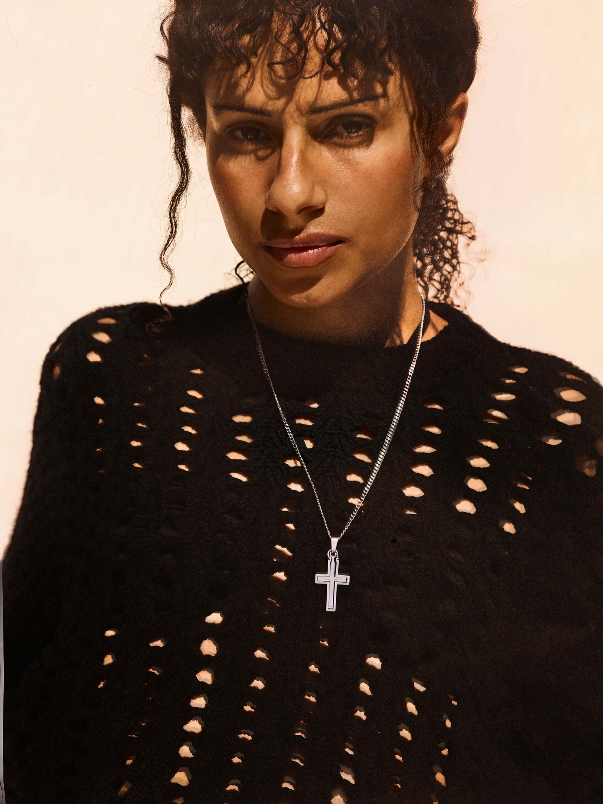 Portrait of a young adult woman with curly hair wearing a black knit sweater and a silver cross necklace against a neutral background.