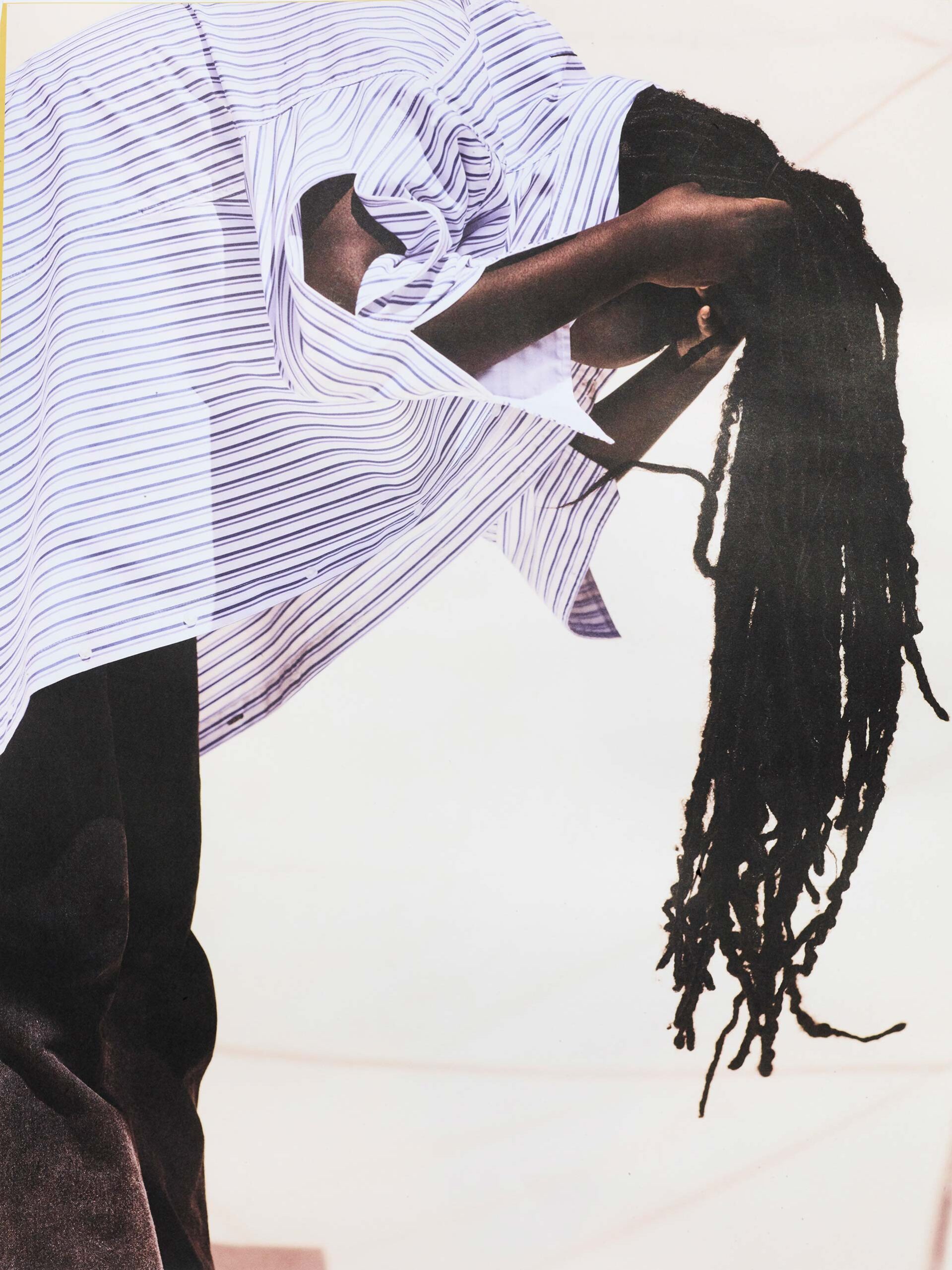 An individual with dreadlocks and a striped shirt leans forward against a pale white background, with soft lighting casting subtle shadows.