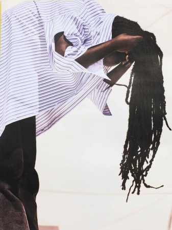 An individual with dreadlocks and a striped shirt leans forward against a pale white background, with soft lighting casting subtle shadows.