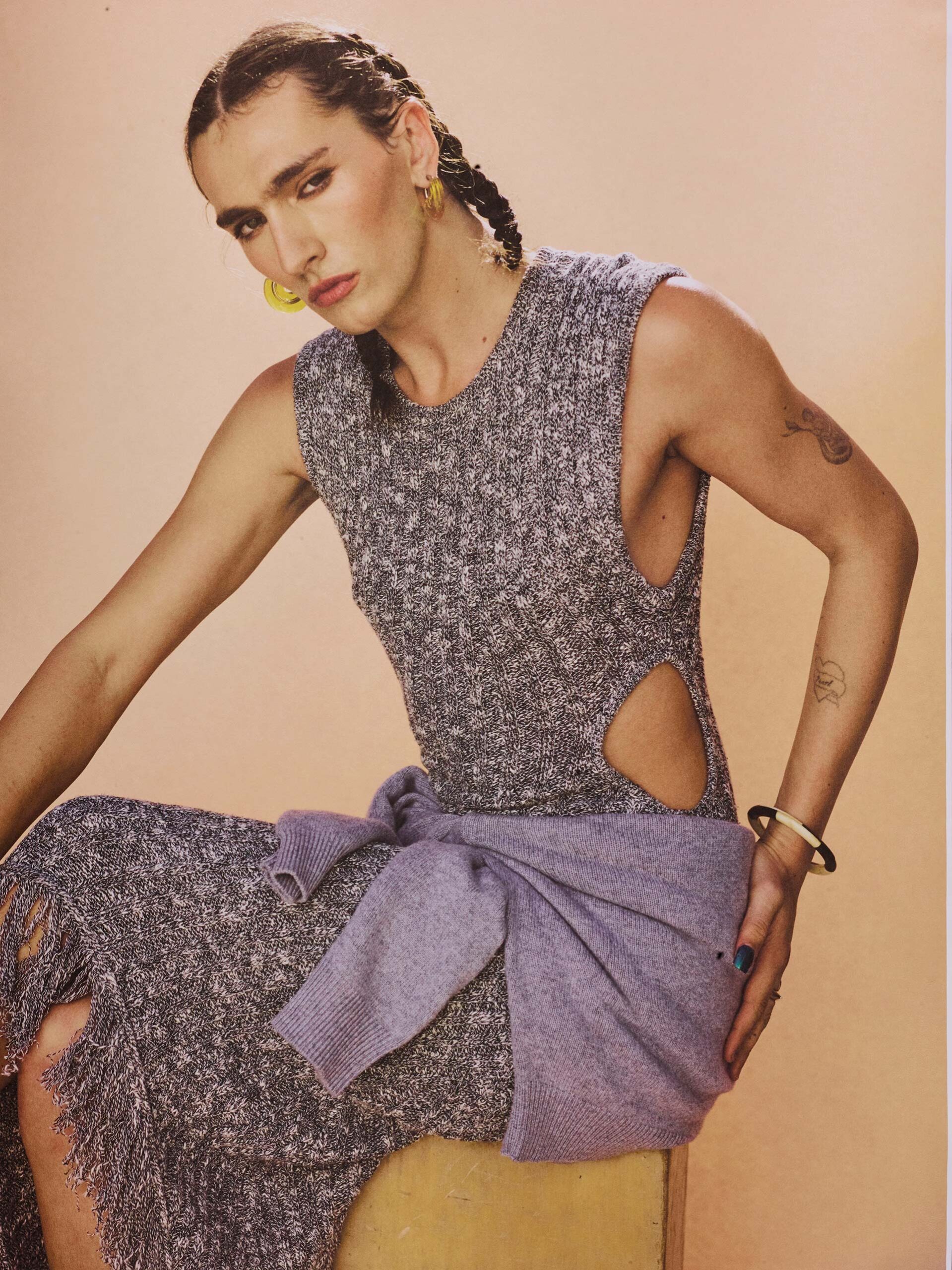 Person with braided hair and bold earrings, wearing a knitted dress with waist tie in front of a gradient background.