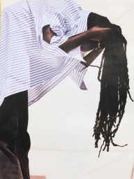 Model with long dreadlocks bending forward wearing a striped button-down shirt and dark pants against a white background