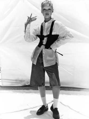 Model in a white shirt and denim shorts wearing a dark harness against a white backdrop