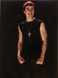 Smiling man with short red hair wearing a black sleeveless top and silver cross necklace against dark background