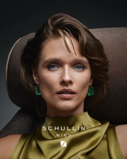 Portrait of a fair-skinned woman with blue eyes wearing green earrings and a satin top, posed in front of a brown armchair.