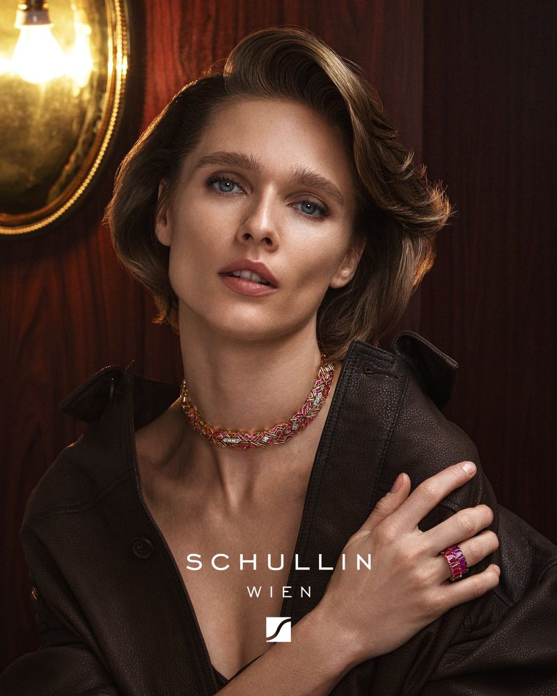 Elegant woman poses wearing a brown leather jacket and jewelry including a ruby ring and necklace inside an indoor setting.