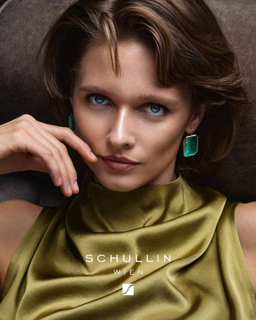Close-up of a woman wearing green emerald earrings, posing in a sleeveless, shiny olive top in a studio. Beauty and fashion.
