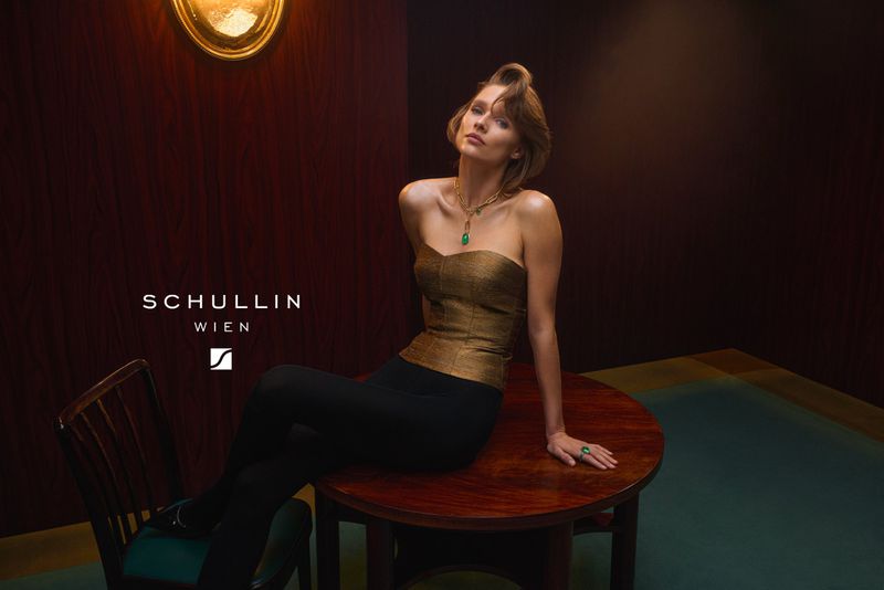 Elegant woman in gold corset and black pants seated at a round wooden table with jewelry and dramatic lighting in an indoor setting.