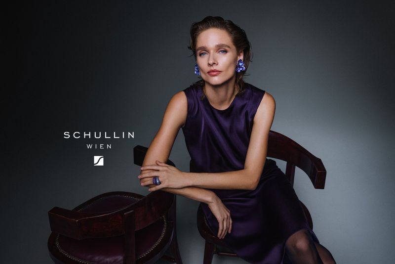 Model seated on a dark wooden chair wearing a purple silk dress and statement earrings against a grey studio backdrop.