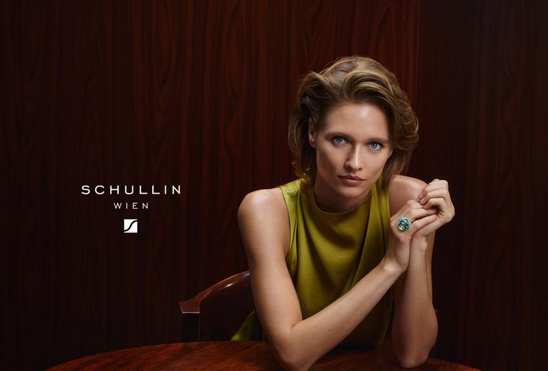 Elegant studio shot of a blond adult woman with a green ring, sitting at a table against a dark brown background, looking at the camera.
