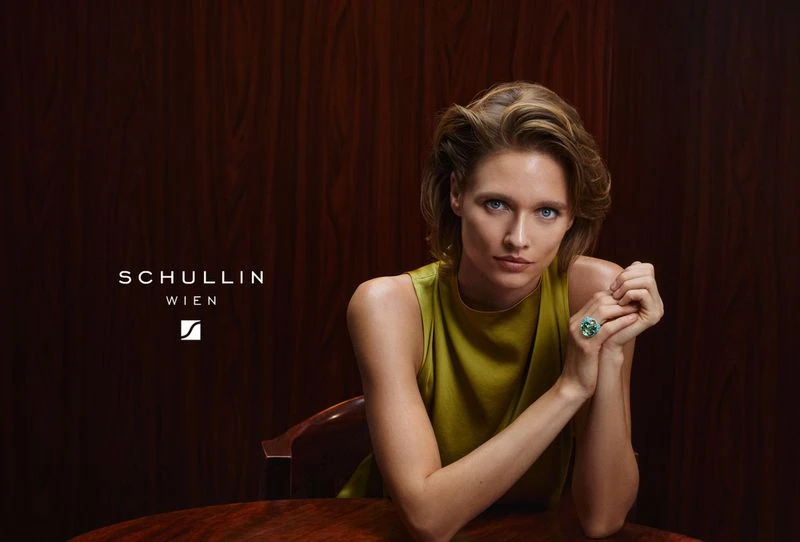 Elegant studio shot of a blond adult woman with a green ring, sitting at a table against a dark brown background, looking at the camera.