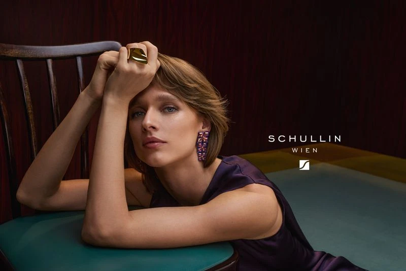 Woman with bob haircut poses indoors wearing large ring and earrings, resting arms on teal chair in front of dark burgundy background.